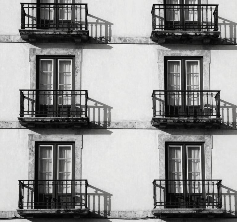 A minimalist architectural detail of a classic Portuguese building facade with clean white stone and dark metal balconies. High contrast between shadows and light, capturing a sense of heritage and quality. Black and white photography.