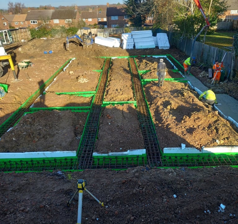Construction site workers using a red concrete pump to pour foundations with steel reinforcement mesh.