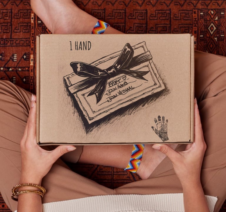 A person holding a 1 Hand cardboard gift box over a patterned rug, showing hand-drawn bow details.