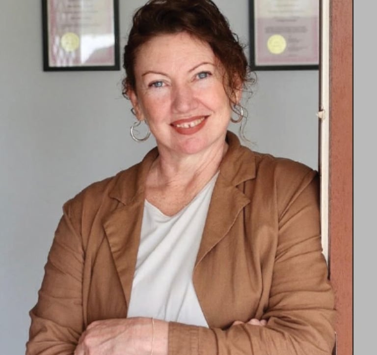 A smiling professional woman with curly dark hair wearing a brown blazer leaning against a doorway.