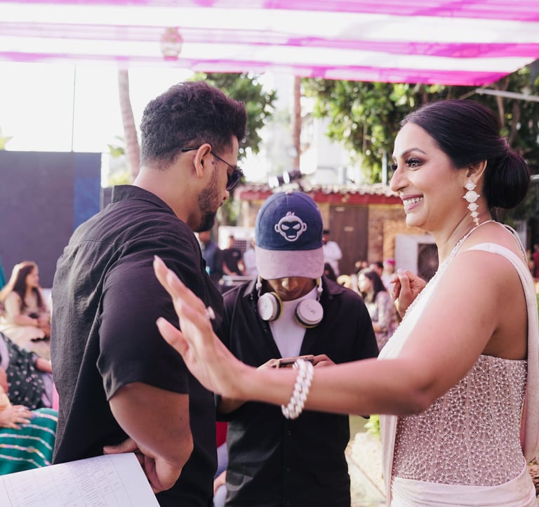 Smiling woman in a beaded dress at an outdoor event with guests and a camera operator.