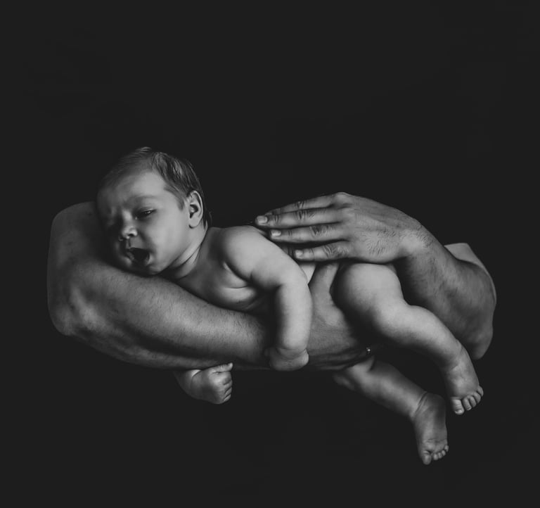 Black and white newborn photography of a father's hands cradling a sleeping baby against a dark background.