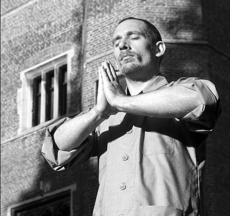 Heng Hu practices mindfulness meditation with hands in prayer position outside a brick building.
