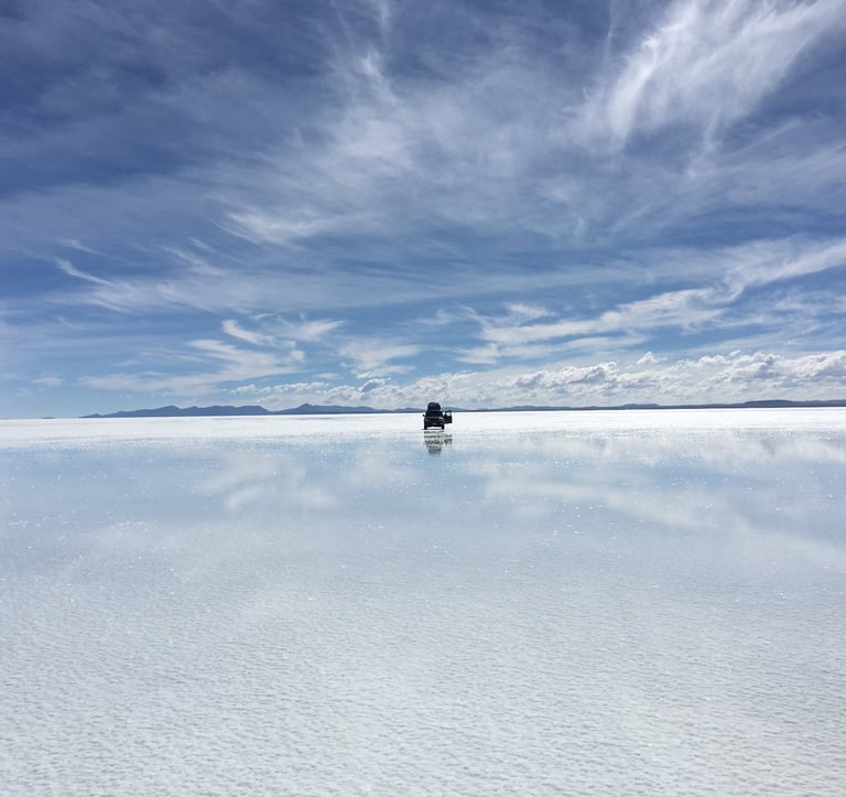 Panorama du Salar d’Uyuni reflétant le ciel bleu avec un 4x4 isolé avançant sur l’eau argentée sous des nuages légers