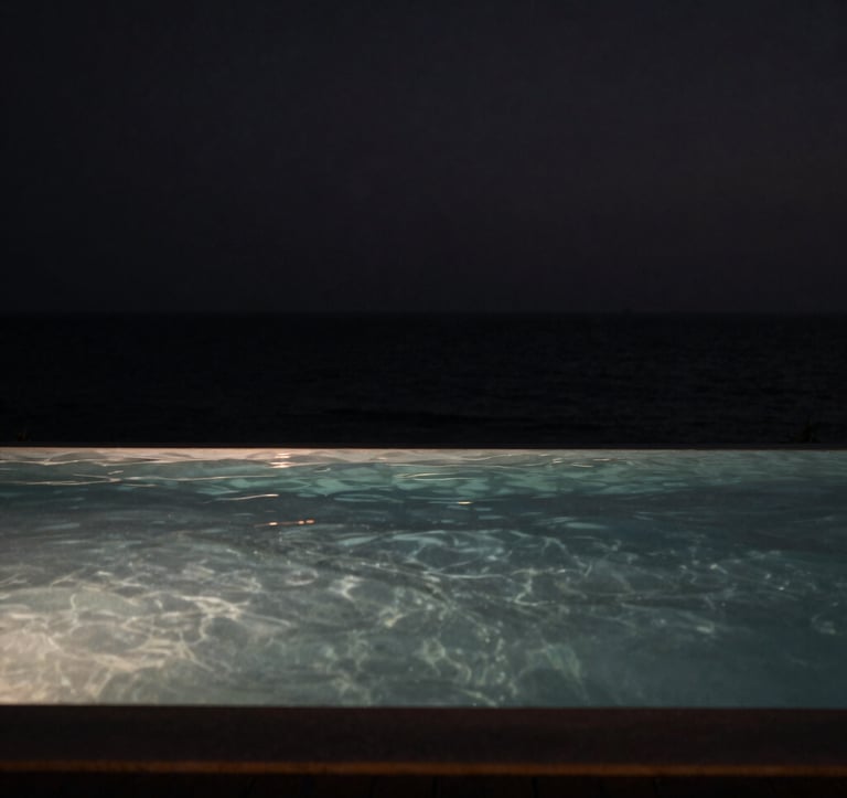 A high-contrast, artistic photography of a tranquil infinity pool meeting the dark ocean horizon at night. The water surface is like glass, reflecting a few pinpricks of soft light. Moody, cinematic atmosphere in shades of black and muted grey. North American / International context.