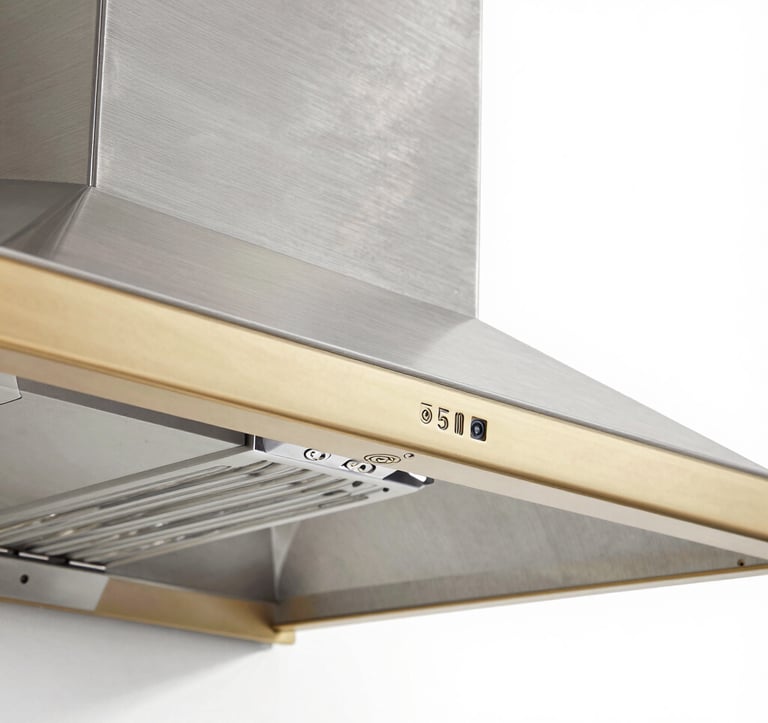 Architectural photography of a technical detail: the corner of a custom range hood where brushed stainless steel meets a champagne gold trim. The focus is on the seamless welding and the precision of the industrial finish, shot in a bright, modern studio setting.