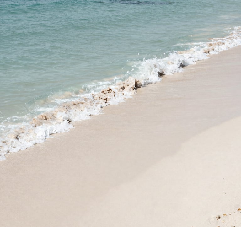 High-angle photography of turquoise ocean waves gently washing over fine white sand on a secluded Brazilian beach. The lighting is bright and clean, capturing the natural beauty of the South American coastline. Colors are dominated by muted blue and off-white.