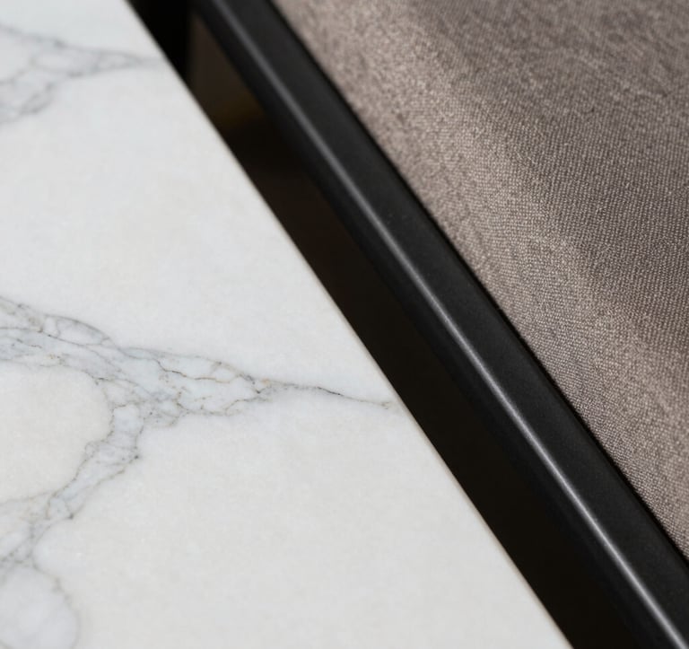 Close-up macro photography of high-end interior textures. A smooth slab of white Indian marble with delicate grey veins sits adjacent to a textured taupe fabric and a matte black metal frame. The lighting is soft and focused, emphasizing the premium quality of the materials.