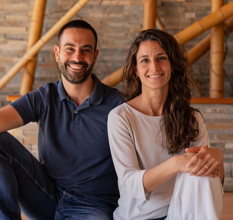 A smiling man and woman sitting together indoors in front of a stone wall and bamboo poles.