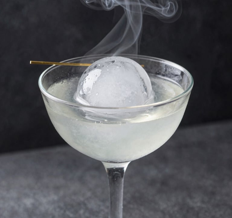 Close-up photography of a transparent cocktail glass featuring a large, clear ice sphere and a minimalist garnish. Wisps of smoke from dry ice drift across a dark charcoal background. North American / Urban.