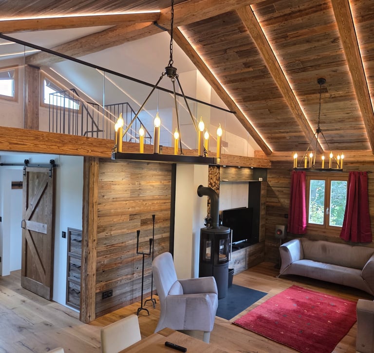 Großzügiges Wohnzimmer mit offenem Altholz-Dachstuhl, Galerie mit Glasgeländer, zwei Kronleuchtern und Kaminofen.