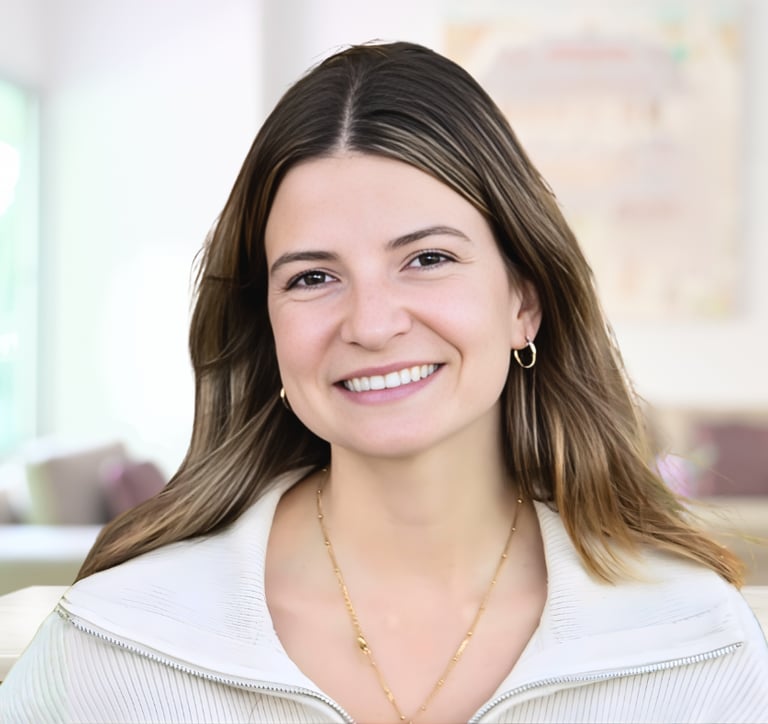 Smiling woman with brown hair and gold jewelry wearing a white sweater in a bright office.