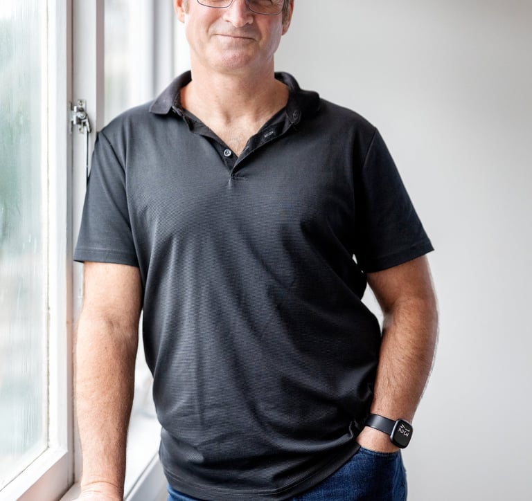 Photo of a man wearing a glasses, a black polo shirt and jeans, leading on a window sill and smiling
