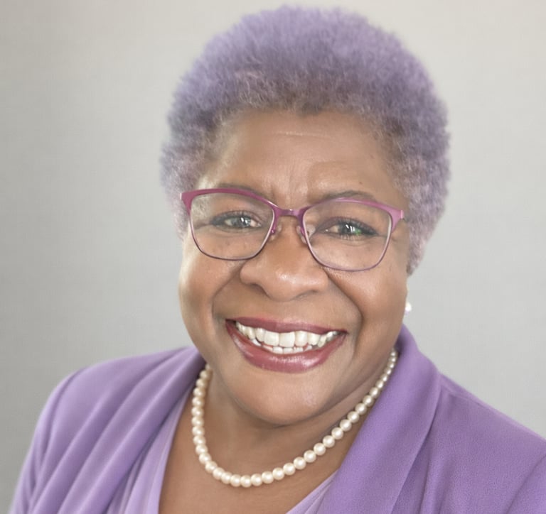 Smiling woman with short purple hair wearing glasses, a pearl necklace, and a purple blazer.