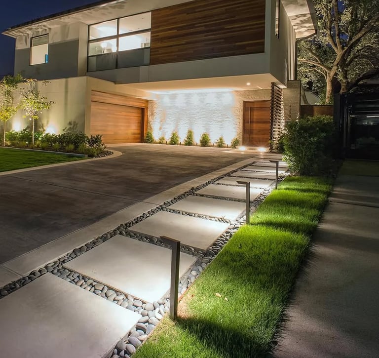 Modern luxury home exterior with illuminated concrete paver walkway and landscape lighting at night.
