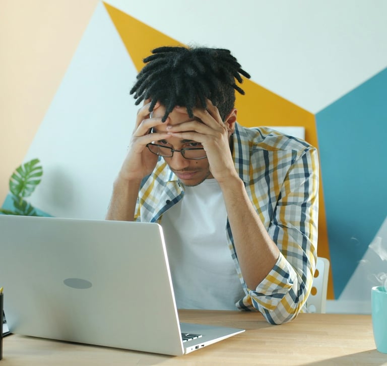 Frustrated young freelancer sitting at a desk with an open  laptop, holding his head in his hands.