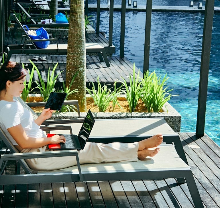 A digital nomad works on a laptop and tablet while lounging by a luxury resort swimming pool.
