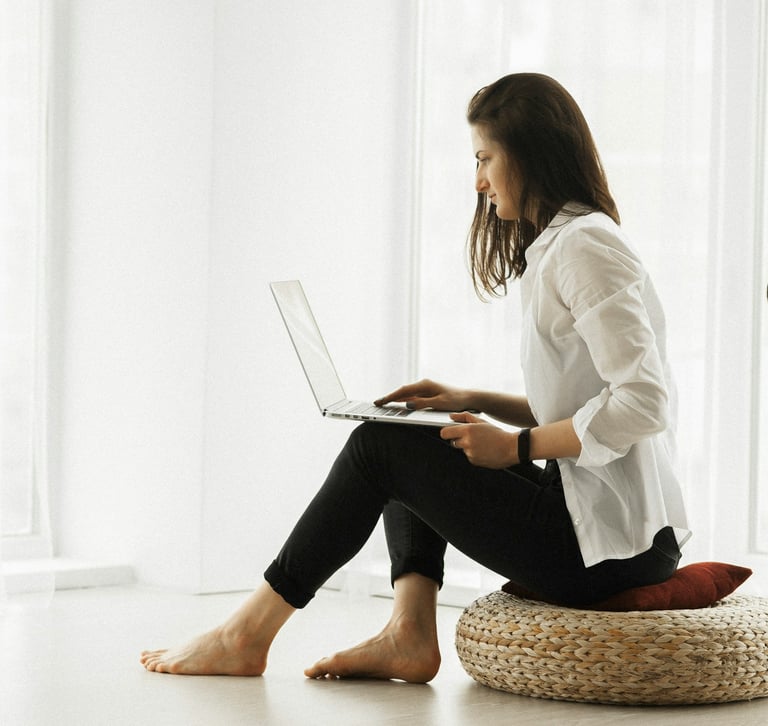 A woman in a white shire and black slacks, sitting on a floor cushion in a white room, working on a laptop.