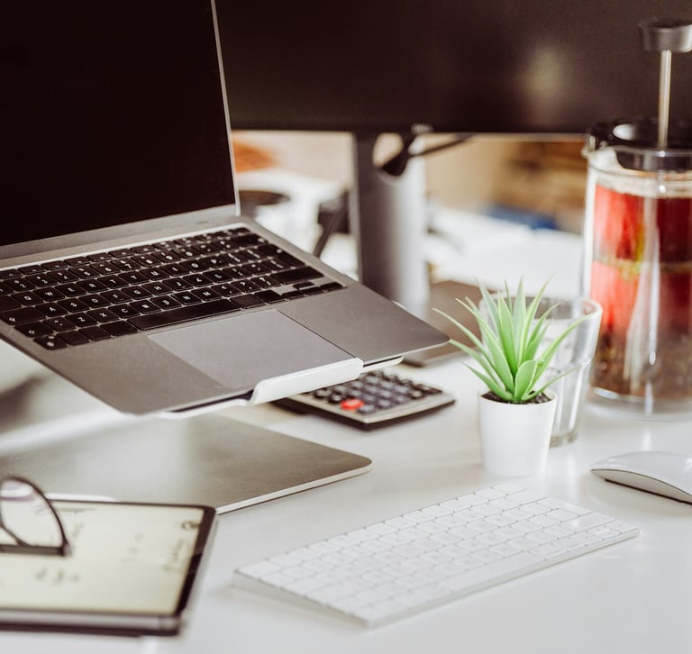 Modern office desk setup with a laptop on a stand, keyboard, mouse, and a French press coffee maker.