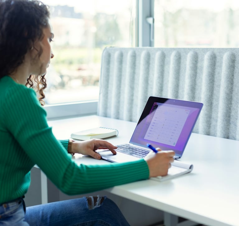 A woman with curly hair works on a laptop at a bright office desk with a notebook and pen..
