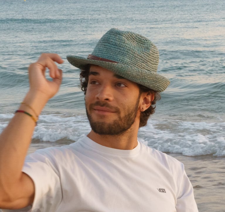 A man wearing a stylish fedora hat and white t-shirt on a sandy beach at sunset.