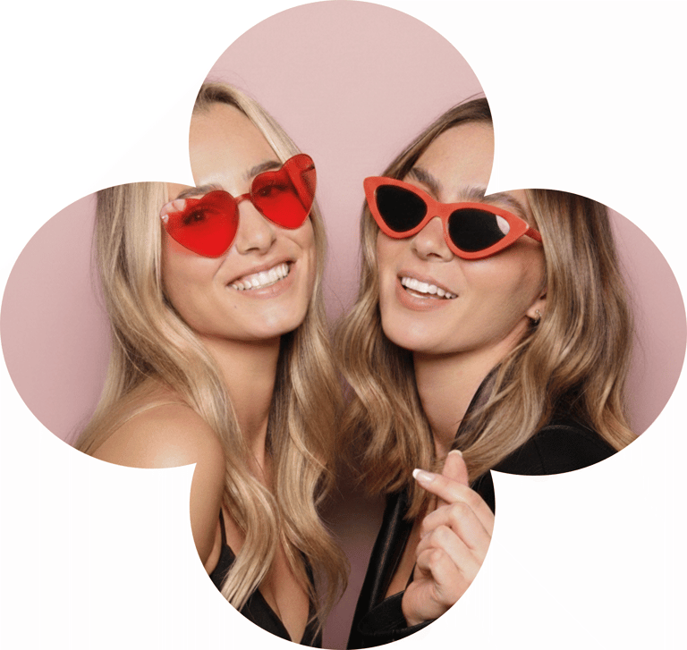 Two smiling women wearing stylish red heart-shaped and cat-eye sunglasses against a pink background.