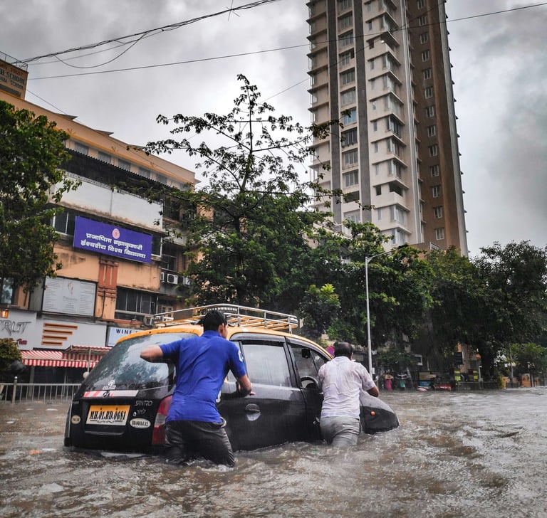 Men pushing a black taxi through deep flood water on a city street in Mumbai during heavy monsoon rain.