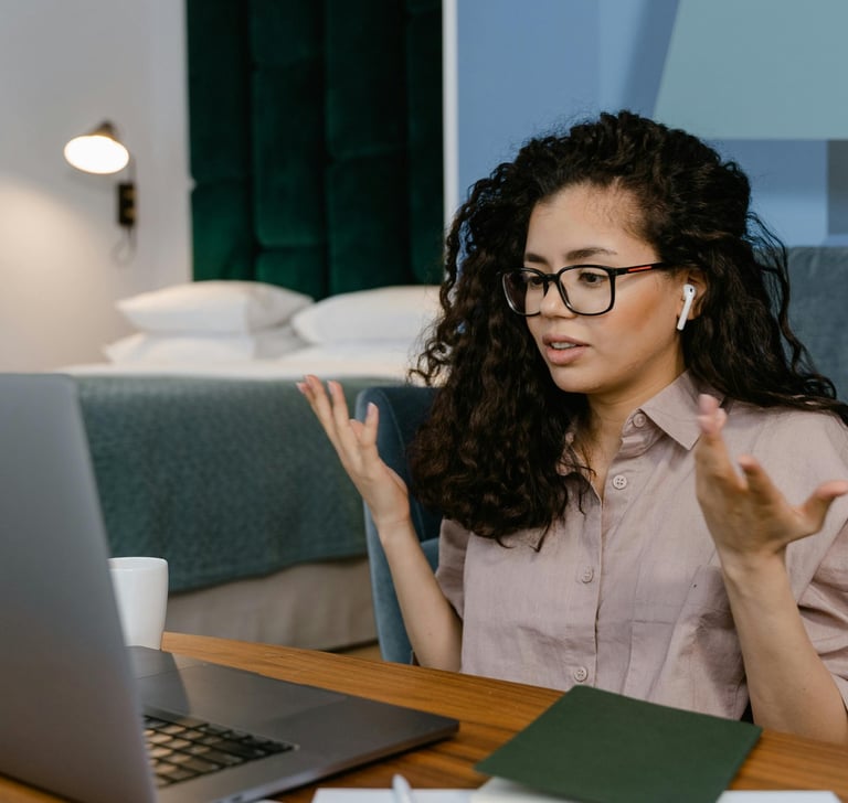 A woman with curly hair wearing glasses joins a video call on her laptop from a home office.