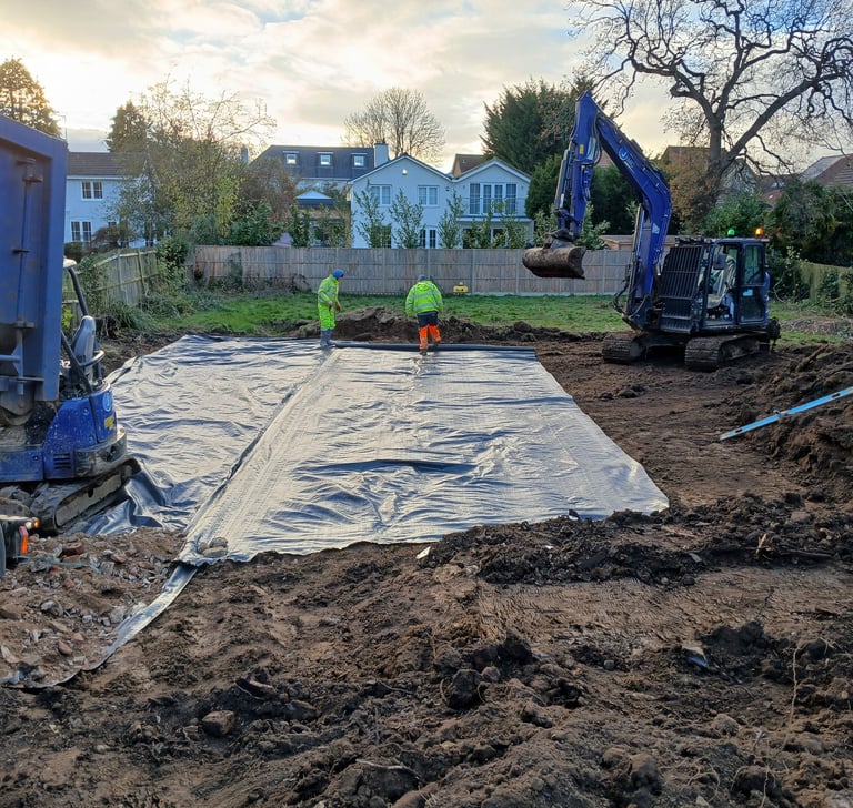 Construction workers laying a damp proof membrane on a residential building site with a blue excavator.