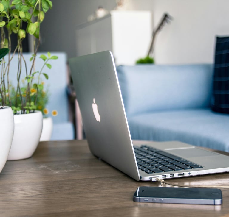 A silver MacBook laptop and iPhone on a wooden coffee table in a modern living room with green plants.