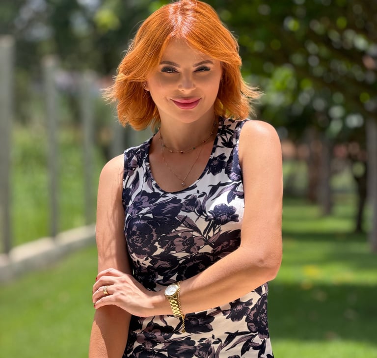 A smiling redhead woman in a floral print sleeveless dress posing outdoors in a sunny park.