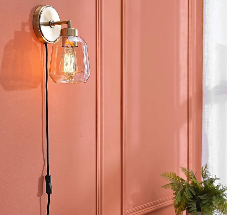 Modern brass wall sconce with a clear glass shade and Edison bulb mounted on a salmon pink wall.