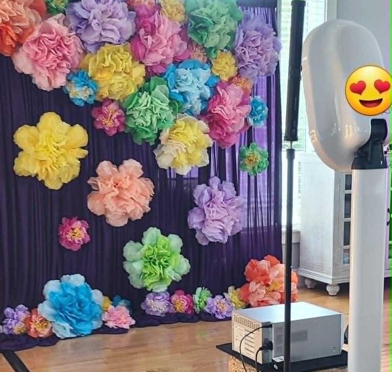 Photo booth setup featuring a purple curtain backdrop adorned with colorful oversized paper flowers.
