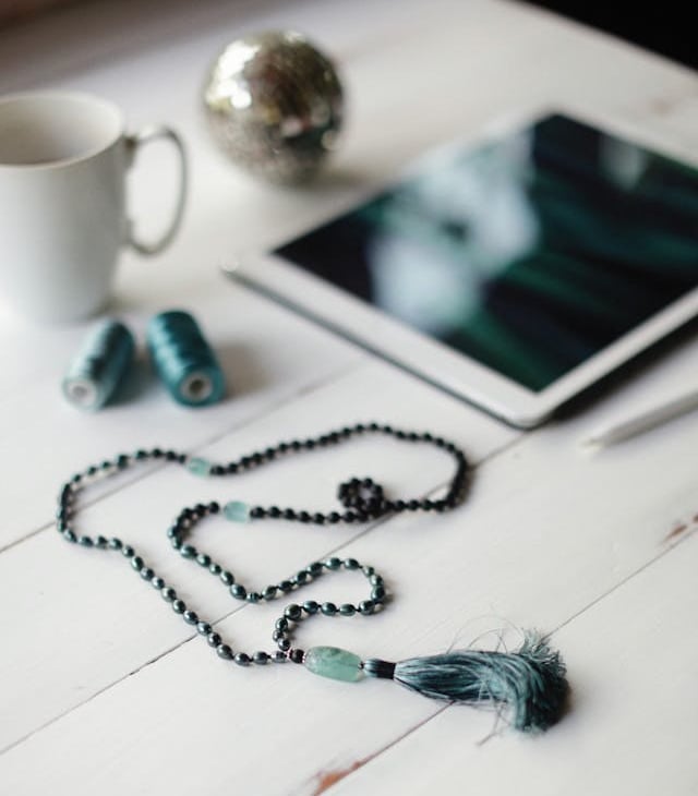 Handmade mala beads with tassel on a white desk next to a digital tablet and coffee mug.