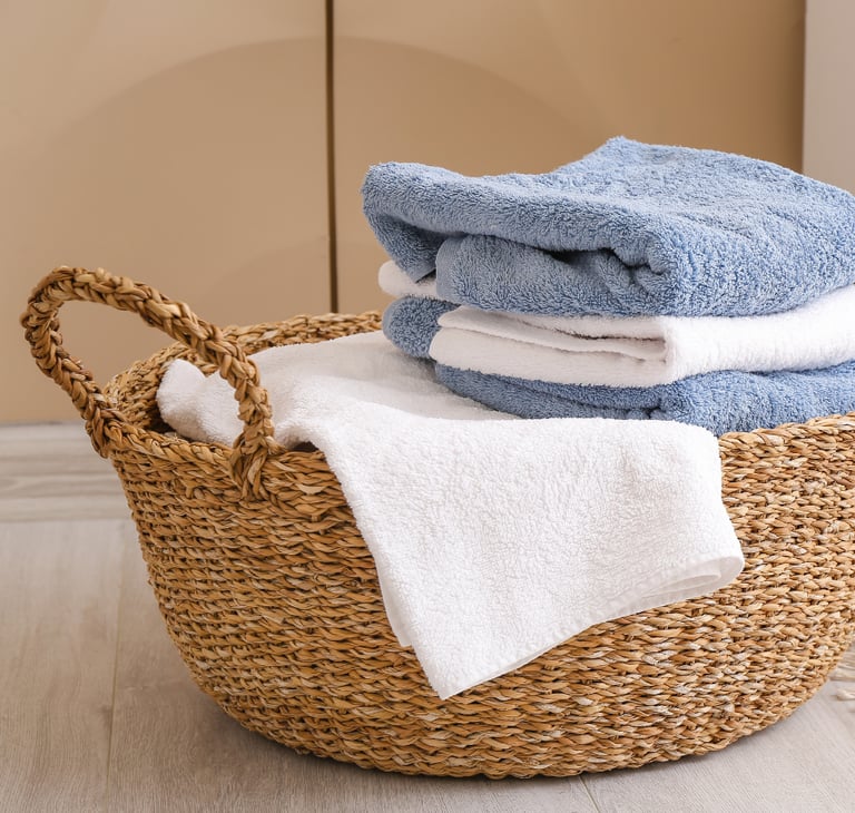 Stack of folded plush blue and white bath towels inside a woven wicker basket on a clean, light hardwood floor. 