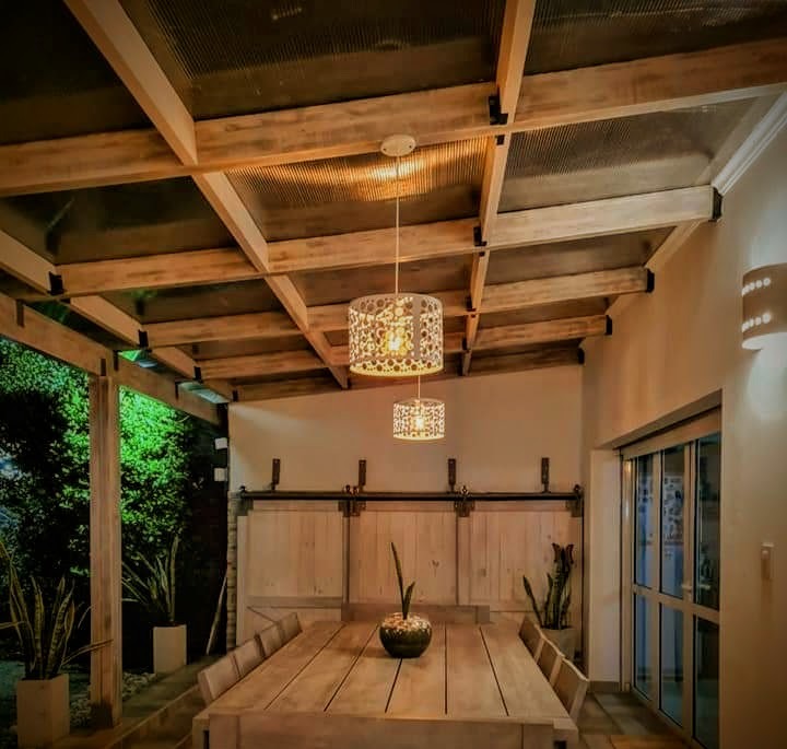 Comedor rústico al aire libre con mesa de madera, iluminación colgante y pérgola de madera.