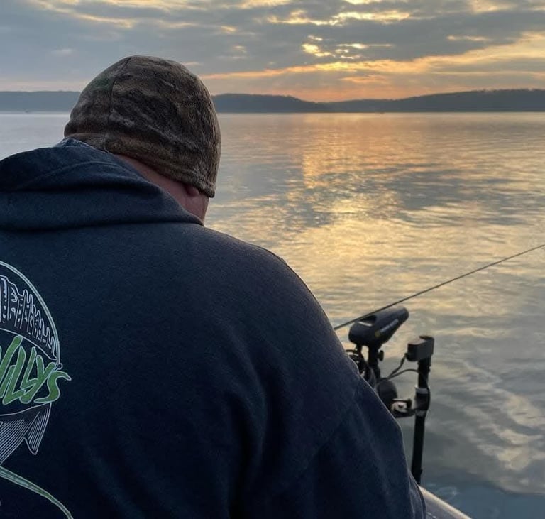A fisherman in a hoodie casts a fishing rod from a boat during a golden sunset on a calm lake.