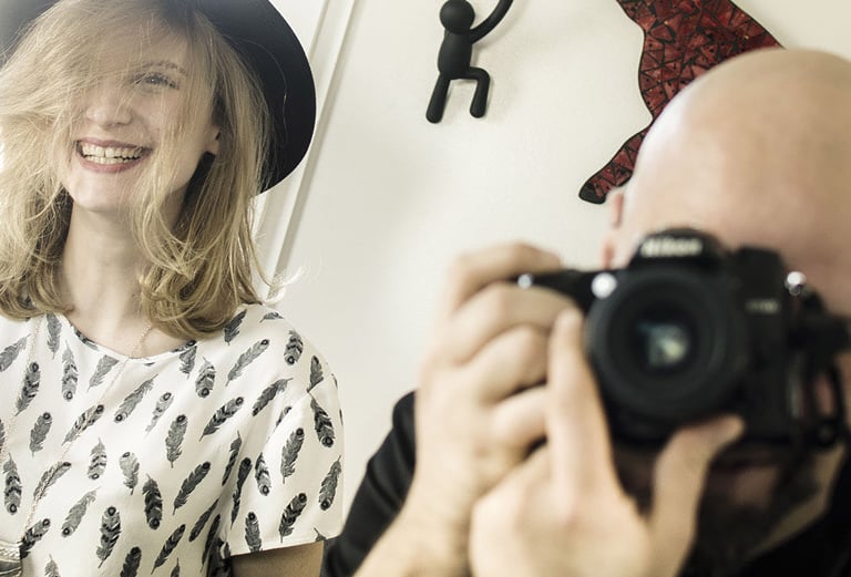A photographer, Séb taking a picture of a smiling woman wearing a black hat and feather print shirt.