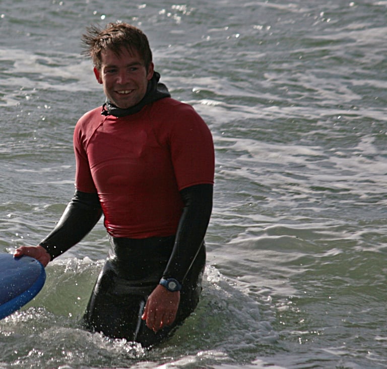 Surf coach Rhys smiling holding the nose of a surfboard in the sea.