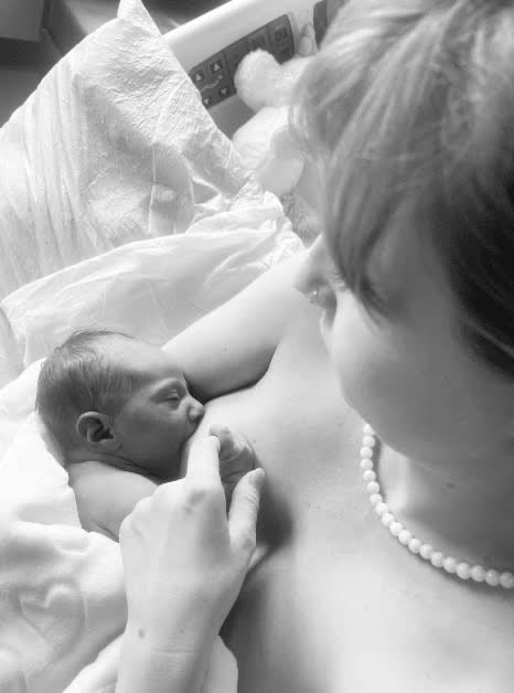 Black and white top view of new mom nursing baby in bed. Baby holds mother's finger as she gazes down with love.