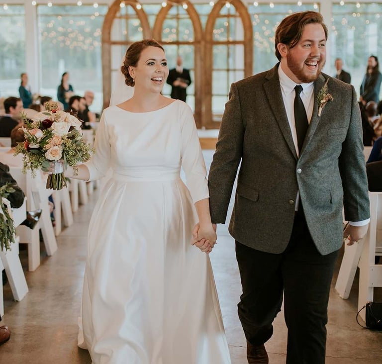 Smiling bride and groom holding hands walking down the aisle in a rustic barn wedding venue.