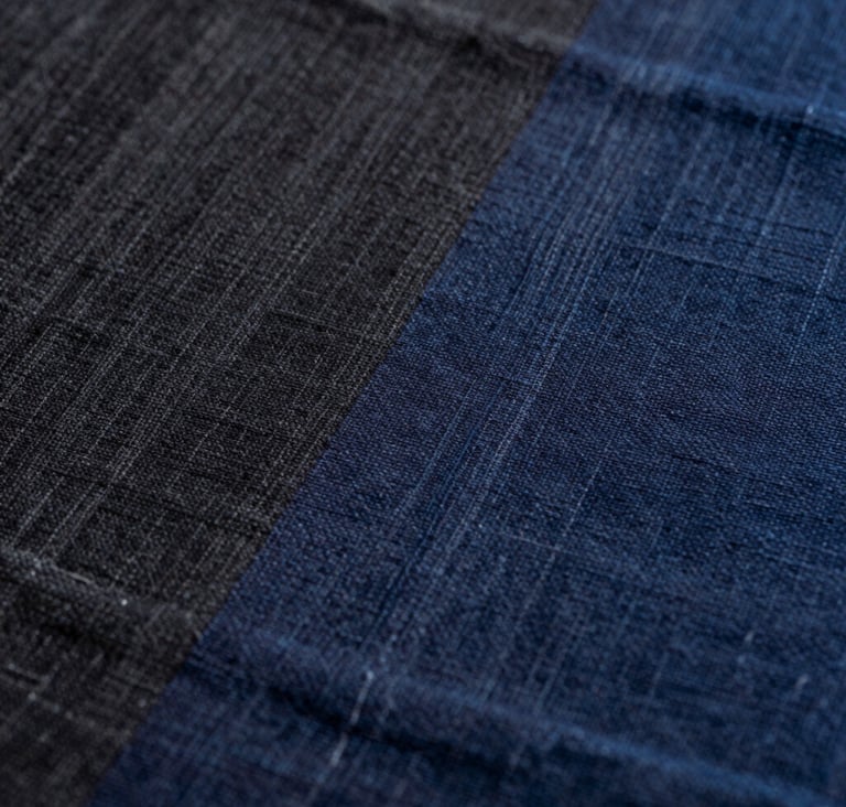 An extreme close-up of textile texture showing the weave of hand-loomed silk. The colors transition from charcoal black to deep indigo, highlighting the organic imperfections of the fiber in a East Asian / South Asian influenced for a global luxury audience design house.