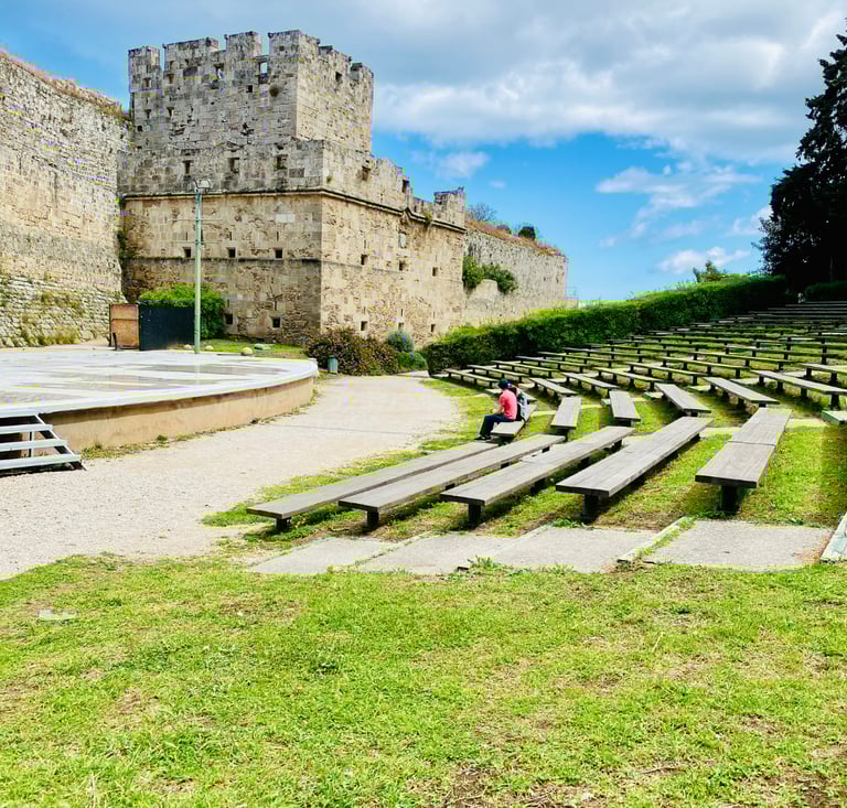 Outdoor stone amphitheater and historic medieval fortress walls in Rhodes, Greece under a blue sky.