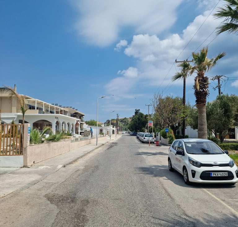 A white Kia parked on a sunny street in Greece with palm trees and Mediterranean architecture.