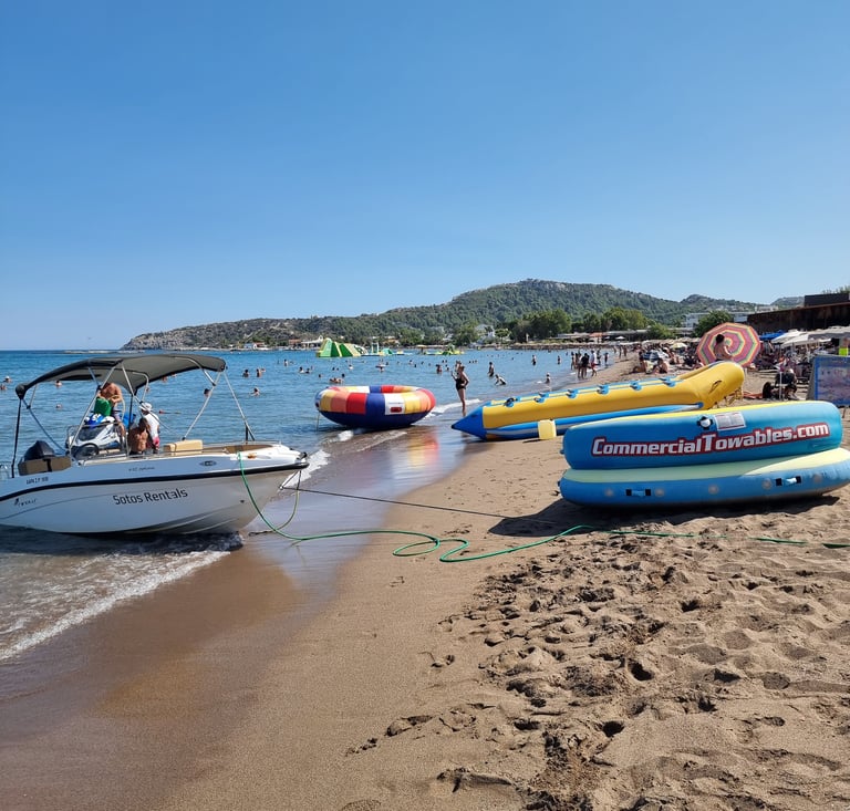 A motorboat and inflatable water sports towables sit on a crowded sandy beach under a clear blue sky.