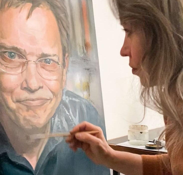 Stefanie placing final touches on oil portrait painting of smiling man with glasses.