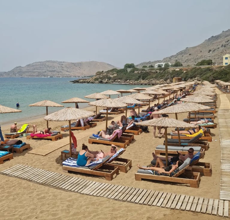 Tourists relaxing on wooden sun loungers under straw umbrellas at a sandy beach resort in Greece.