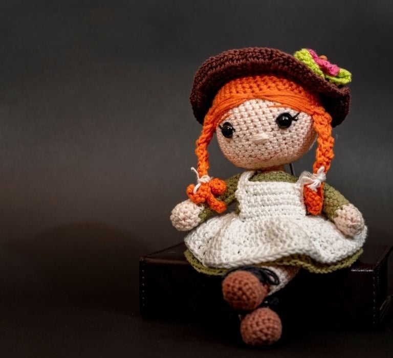 Muñeca tejida a crochet hecha a mano con trenzas rojas y vestido vintage sentada sobre una caja