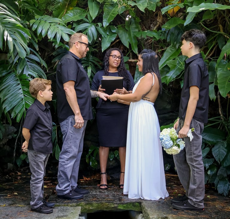 A couple exchanges vows at an outdoor tropical garden wedding ceremony with an officiant and children.