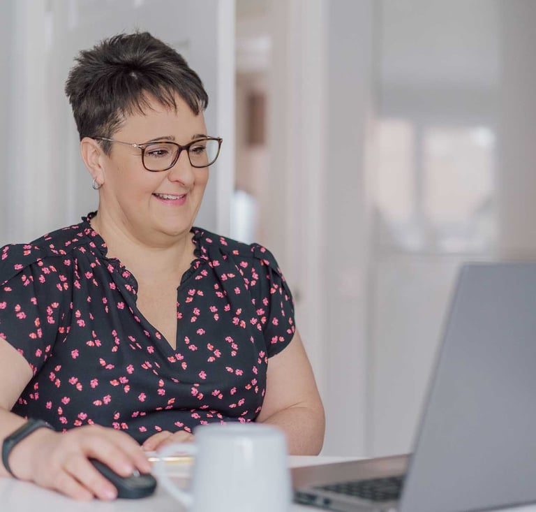 Smiling woman with short hair and glasses working on her laptop at a desk.
